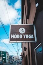 A street view features a wooden sign for 'Modo Yoga' with a flame logo, hanging from a building. The background reveals an urban setting with colorful buildings, power lines, and a partly cloudy sky.