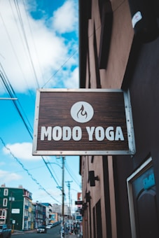 A street view features a wooden sign for 'Modo Yoga' with a flame logo, hanging from a building. The background reveals an urban setting with colorful buildings, power lines, and a partly cloudy sky.