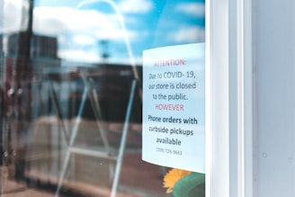 The image shows a sign posted on a glass door, indicating that the store is closed to the public due to COVID-19 but offering phone orders with curbside pickups. The background appears blurred, possibly a street or urban area.