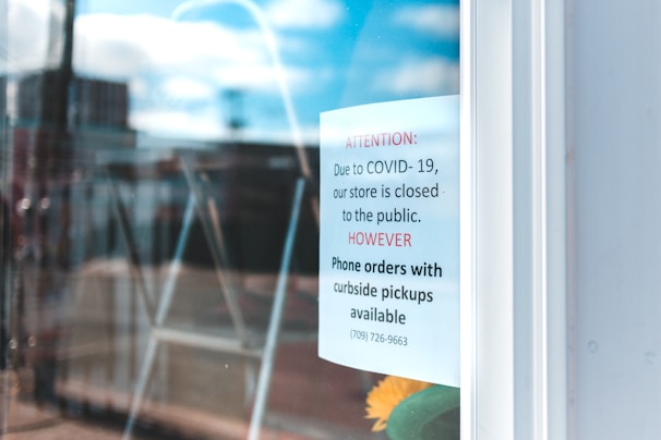The image shows a sign posted on a glass door, indicating that the store is closed to the public due to COVID-19 but offering phone orders with curbside pickups. The background appears blurred, possibly a street or urban area.