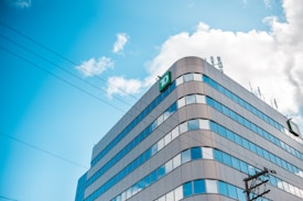 A modern office building with a distinctive green 'TD' logo on its corner, featuring a glass facade reflecting the blue sky and scattered clouds. Telephone lines are visible in the foreground, while several antennas are situated on the roof.
