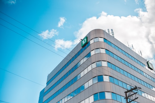 A modern office building with a distinctive green 'TD' logo on its corner, featuring a glass facade reflecting the blue sky and scattered clouds. Telephone lines are visible in the foreground, while several antennas are situated on the roof.