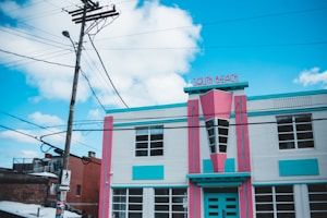 An art deco style building adorned with vibrant pink and turquoise colors. The facade features geometric patterns, large windows, and a sign that reads 'South Beach'. Surrounding the building are utility poles with electrical wires stretching across the sky. The sky is bright with scattered clouds, and the street around the building is partially visible.