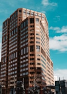 A tall, brown modern office building with numerous windows stands under a bright blue sky with scattered clouds. Below, there are trees without leaves and some street elements like signs and partial views of other buildings. The architecture features a mix of rectangular shapes and angular facets, giving it a slightly geometric look.