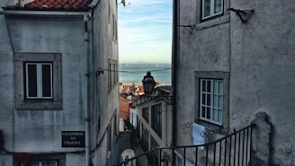 A narrow street flanked by old buildings with white and grey facades. There is a glimpse of the sea in the distance through the gap between the structures. A traditional street lamp hangs in the center, and a visible street sign reads 'Calcadinha da Figueira.' The image portrays a typical coastal urban setting.