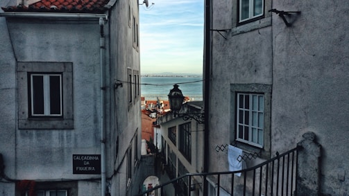 A narrow street flanked by old buildings with white and grey facades. There is a glimpse of the sea in the distance through the gap between the structures. A traditional street lamp hangs in the center, and a visible street sign reads 'Calcadinha da Figueira.' The image portrays a typical coastal urban setting.