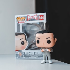 A Pop! Vinyl figure resembling a character with dark hair and a gray suit stands in front of a matching box. The box has a cartoon portrayal of the character similar to the figure and displays the number 644 and the name of the character, along with the word 'Fun'. The figure and box are placed on a light-colored surface with a dark background.