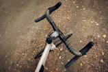 A bicycle with drop handlebars is positioned on a dirt path covered with scattered leaves. The handlebars are wrapped in tape, and there is a small electronic device mounted at the center of the handlebars.