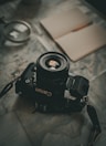 A vintage film camera rests on a detailed map, accompanied by an open notebook and magnifying glass. The setting suggests exploration or travel planning, with muted lighting creating a nostalgic ambiance.