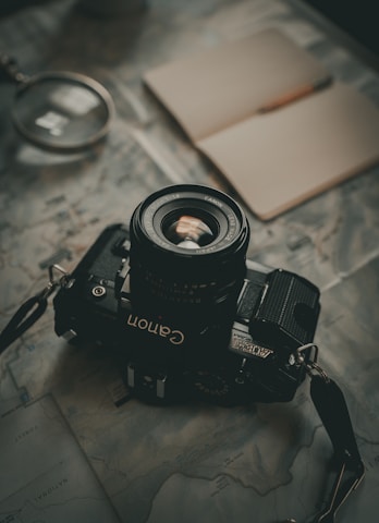 A vintage film camera rests on a detailed map, accompanied by an open notebook and magnifying glass. The setting suggests exploration or travel planning, with muted lighting creating a nostalgic ambiance.
