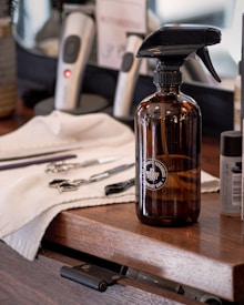 A barbershop countertop with a brown spray bottle labeled '85 Bergen Street' placed prominently in the foreground. Behind the bottle are grooming tools including scissors, combs, and clippers, some of which are resting on a white towel. The countertop itself is made of wood with a slightly open drawer visible at the bottom.