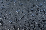 Water droplets of various sizes scattered across a smooth surface. The droplets create a textured pattern against a dark background, reflecting light and giving a glossy appearance.