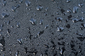Water droplets of various sizes scattered across a smooth surface. The droplets create a textured pattern against a dark background, reflecting light and giving a glossy appearance.