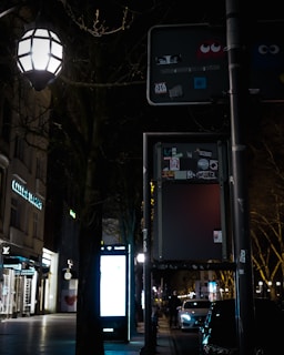 A dimly lit urban street scene features a vintage-style street lamp illuminating the area, casting light on a nearby signpost covered in stickers, including a Pac-Man icon. The background showcases a row of closed shops with branded storefronts and a digital billboard glowing in the night, while parked cars line the street.