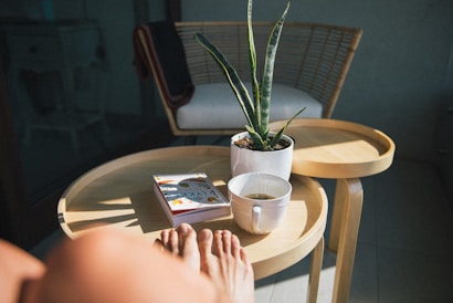 A cozy setting with sunlight streaming onto a wooden side table, which holds a potted plant, a book, and a cup of tea. In the foreground, a person's feet are visible, suggesting relaxation and leisure. In the background, a wicker chair with a cushion is partially visible.