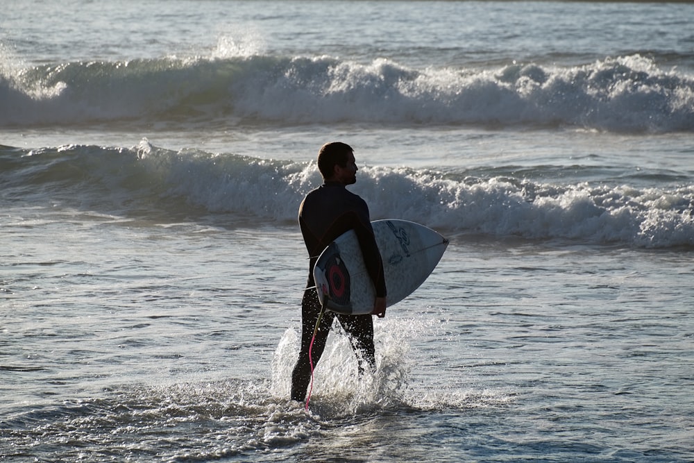 homem na camisa preta segurando a prancha de surf branca nas ondas do mar durante o dia