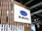 A large sign featuring the Subaru logo, a blue oval with stars, is mounted on a wooden panel with vertical slats. The scene appears to be indoors, possibly at a car showroom or event space, with industrial ceiling elements and lighting fixtures visible in the background.