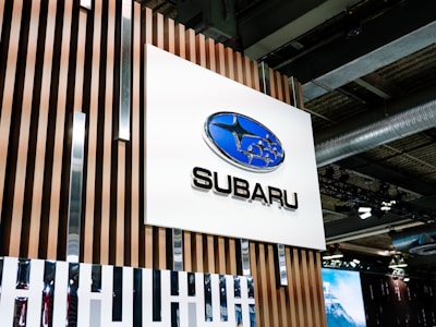 A large sign featuring the Subaru logo, a blue oval with stars, is mounted on a wooden panel with vertical slats. The scene appears to be indoors, possibly at a car showroom or event space, with industrial ceiling elements and lighting fixtures visible in the background.