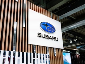 A large sign featuring the Subaru logo, a blue oval with stars, is mounted on a wooden panel with vertical slats. The scene appears to be indoors, possibly at a car showroom or event space, with industrial ceiling elements and lighting fixtures visible in the background.