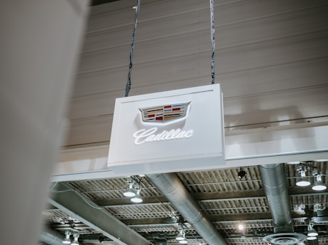 A well-lit indoor space with overhead industrial lighting and visible ductwork. A white sign with the Cadillac logo and name hangs from the ceiling.