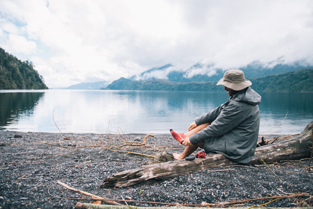 man in gray jacket sitting on brown tree log near body of water during daytime