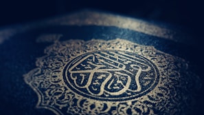 Intricate Arabic calligraphy and ornate patterns are embossed in gold on a dark background, creating a sense of elegance and reverence.