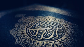 Intricate Arabic calligraphy and ornate patterns are embossed in gold on a dark background, creating a sense of elegance and reverence.