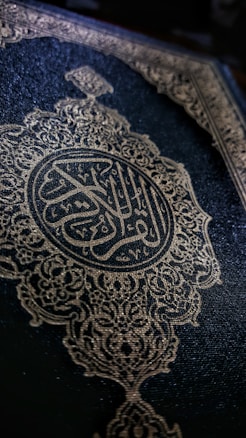 An ornate book cover featuring intricate gold arabesque designs and Arabic calligraphy. The dark background accentuates the elaborate patterns, conveying a sense of tradition and reverence.