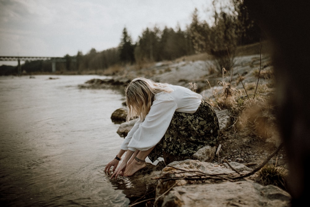 woman in white long sleeve shirt sitting on brown rock near body of water during daytime