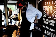 A person wearing a hoodie and a floral hat is adjusting their apron in what appears to be a tattoo parlor. The surroundings include tattoo equipment and a large sign on the wall with text related to tattoo services.
