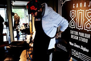 A person wearing a hoodie and a floral hat is adjusting their apron in what appears to be a tattoo parlor. The surroundings include tattoo equipment and a large sign on the wall with text related to tattoo services.