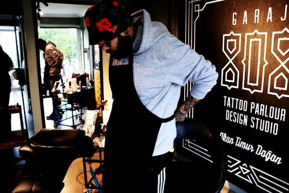 A person wearing a hoodie and a floral hat is adjusting their apron in what appears to be a tattoo parlor. The surroundings include tattoo equipment and a large sign on the wall with text related to tattoo services.