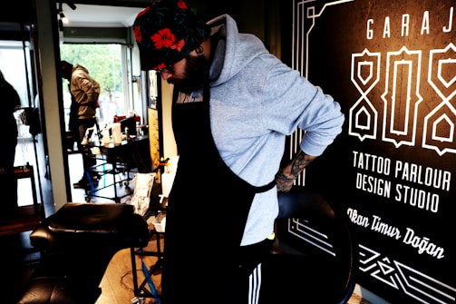 A person wearing a hoodie and a floral hat is adjusting their apron in what appears to be a tattoo parlor. The surroundings include tattoo equipment and a large sign on the wall with text related to tattoo services.