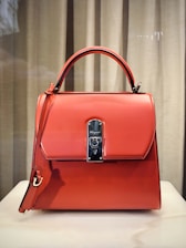 A stylish, orange leather handbag with a structured design is placed against a neutral backdrop. It features a prominent metal clasp in the center, a top handle, and an additional strap hanging down the side.