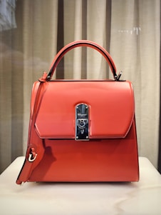 A stylish, orange leather handbag with a structured design is placed against a neutral backdrop. It features a prominent metal clasp in the center, a top handle, and an additional strap hanging down the side.