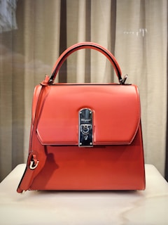 A stylish, orange leather handbag with a structured design is placed against a neutral backdrop. It features a prominent metal clasp in the center, a top handle, and an additional strap hanging down the side.