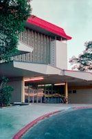 A modern architectural building with a distinctive red roof and vertical column design stands amidst greenery. The entrance features tall glass windows with abstract patterns. A few bicycles are parked under the shelter of the entrance canopy, suggesting a university or communal space. The scene is serene, with no people present.