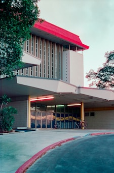 A modern architectural building with a distinctive red roof and vertical column design stands amidst greenery. The entrance features tall glass windows with abstract patterns. A few bicycles are parked under the shelter of the entrance canopy, suggesting a university or communal space. The scene is serene, with no people present.