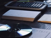 A wooden desk holds a calculator, stacks of paper, and a CD. Papers are organized and placed across the surface, while the CD reflects light with its iridescent surface.