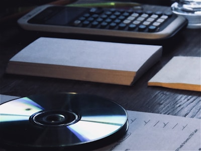 A wooden desk holds a calculator, stacks of paper, and a CD. Papers are organized and placed across the surface, while the CD reflects light with its iridescent surface.