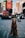 A golden retriever wearing a harness is sitting on a busy city street. The background features blurred lights and advertisements, resembling an urban area like Times Square. The dog appears calm and friendly.