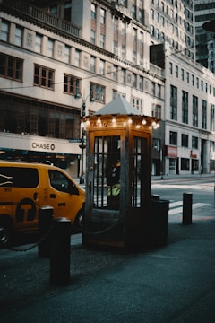 A vintage-looking taxi stand booth illuminated with soft yellow lights is situated on a city street. Nearby, a classic yellow New York City taxi is parked, partially obscured by dark bollards that are connected with chains. Tall, historic buildings with a mix of old and modern architectural styles line the street, creating an urban backdrop.