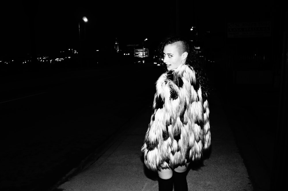 grayscale photo of woman in fur coat standing on road
