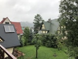 A scenic view of several houses with pitched roofs surrounded by lush greenery. The garden area is bordered by a variety of trees, and one of the roofs features solar panels. In the foreground, a hammock is hung and a small fire pit is visible, adding to the cozy atmosphere.