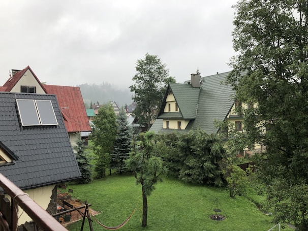A scenic view of several houses with pitched roofs surrounded by lush greenery. The garden area is bordered by a variety of trees, and one of the roofs features solar panels. In the foreground, a hammock is hung and a small fire pit is visible, adding to the cozy atmosphere.