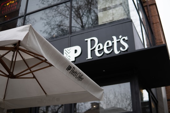 The front of a Peet's Coffee shop is seen with a large, black and white sign above the entrance. In the foreground, a white umbrella featuring the Peet's Coffee logo partially blocks the view of the building. The structure has large glass windows with reflections and bare tree branches visible.