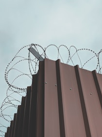 A tall metal fence with sharp-edged vertical panels topped with barbed wire spirals that curl into the sky, creating a sense of security and enclosure. The sky above is overcast, giving the scene a muted and stark atmosphere.