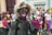 A person is dressed in a dramatic costume with dark body paint and bright pink accents, including gloves with long, claw-like nails. The individual has a wild, blonde wig and is in an expressive pose. A crowd of people, including children, are in the background, suggesting a festive event.