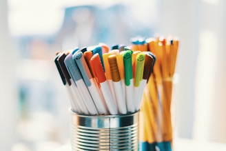 Various colored pens are placed in a tin can, with another set of pencils standing upright in the background. The pens have caps in multiple colors, including orange, blue, green, and yellow. The scene is brightly lit, possibly by natural light, creating a relaxed and creative atmosphere.