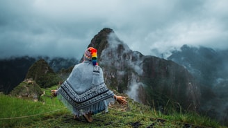 A person wrapped in a colorful woven shawl sits in a meditative pose on a grassy slope overlooking a misty, mountainous landscape. The person faces away, revealing a vibrant knitted hat with multi-colored tassels. Clouds partially obscure the steep cliffs and lush greenery in the background, creating a mystical and serene atmosphere.